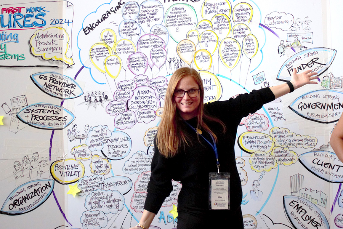 A woman with long red hair and glasses stands smiling in front of a large, colorful graphic recording wall filled with hand-drawn illustrations, text, and diagrams.