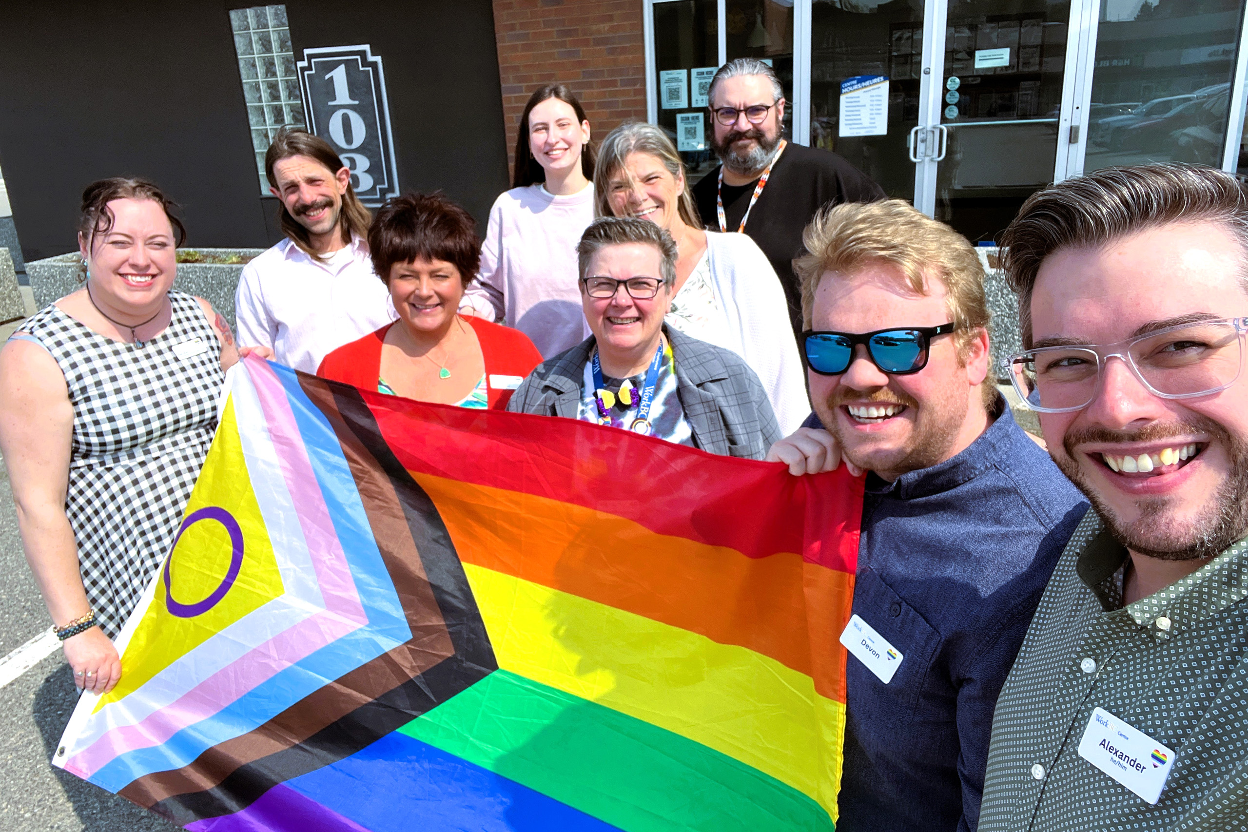 WCG employees smile posing together outdoors, holding a Progress Pride flag that includes the intersex symbol.
