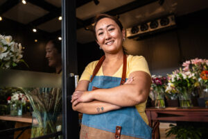 A smiling woman wearing an apron stands with arms crossed in a flower shop.